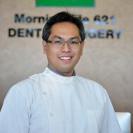 Smiling Asian man, likely a dentist, wearing glasses and a white coat, in front of