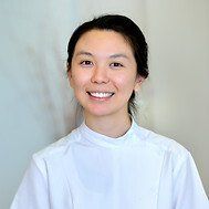 Woman with dark hair pulled back, smiling, wearing a white collared shirt, against a neutral background.