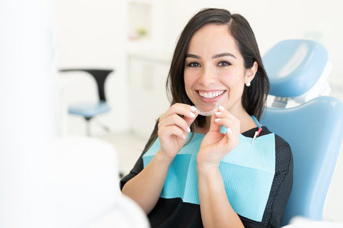 Woman in a dentist's chair smiles, holding clear aligners. She wears a bib and is in a bright dental office.