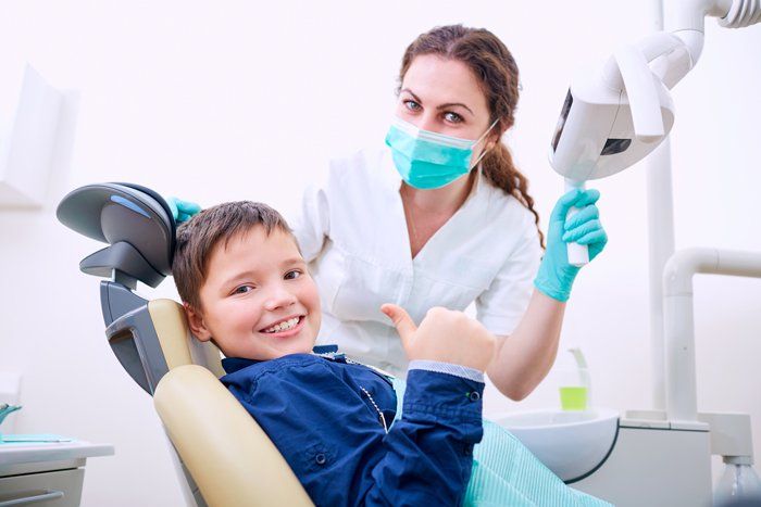 Boy in a dentist chair giving a thumbs up, with dentist in mask and gloves holding the exam light.