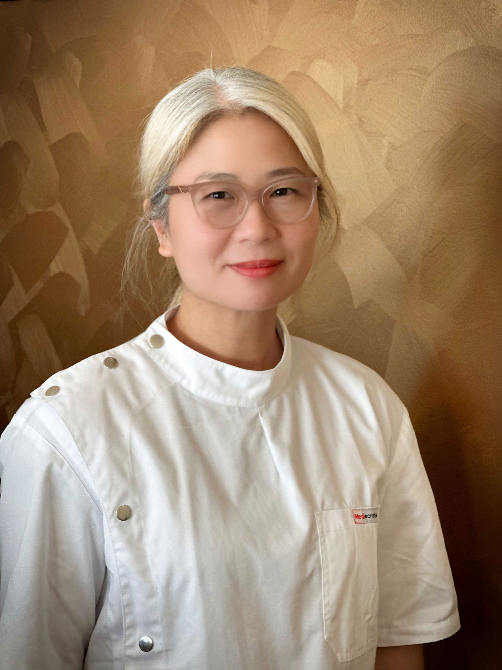 Woman with glasses and silver hair in a white uniform, smiling slightly in front of a textured gold background.