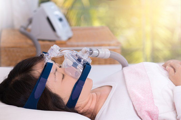 Woman asleep in bed wearing a CPAP mask connected to a machine. The setting is a brightly lit room.